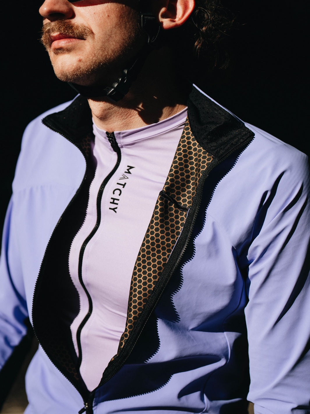 Coupe-vent vélo MATCHY coloris lavande porté par un cycliste équipé d'un casque blanc et lunettes de soleil, zip central et poignets ajustés.