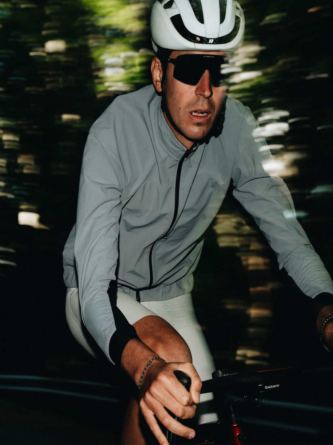 Coupe-vent vélo MATCHY coloris gris porté par un cycliste équipé d'un casque blanc et lunettes de soleil, zip central et poignets ajustés.